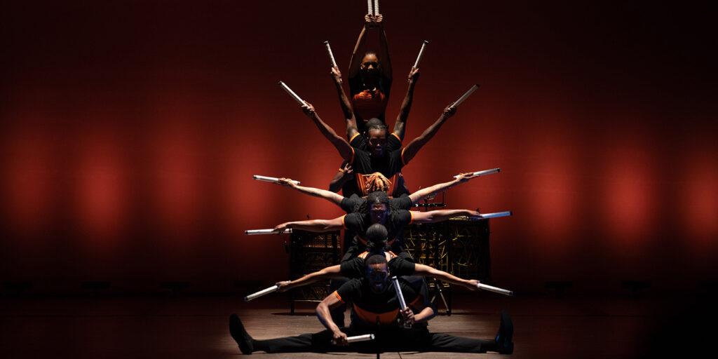 A group of dancers in black clothing holds arms outstretched while on a stage in front of a red backdrop.