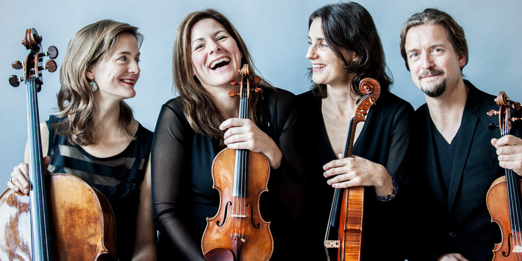 Four musicians in black clothing hold instruments in front of a pale blue wall.