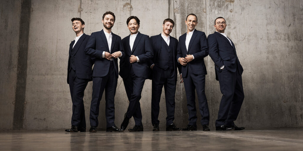 Six men in dark suits and white shirts stand in front of a gray concrete wall.