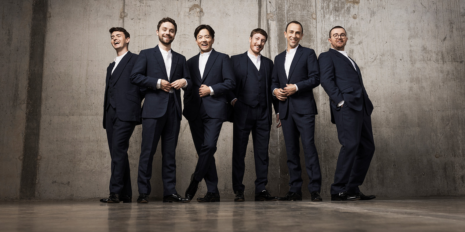 Six men in dark suits and white shirts stand in front of a gray concrete wall.