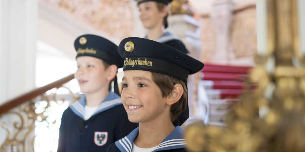 Three boys in uniforms sing while on the stairs of a Baroque palace