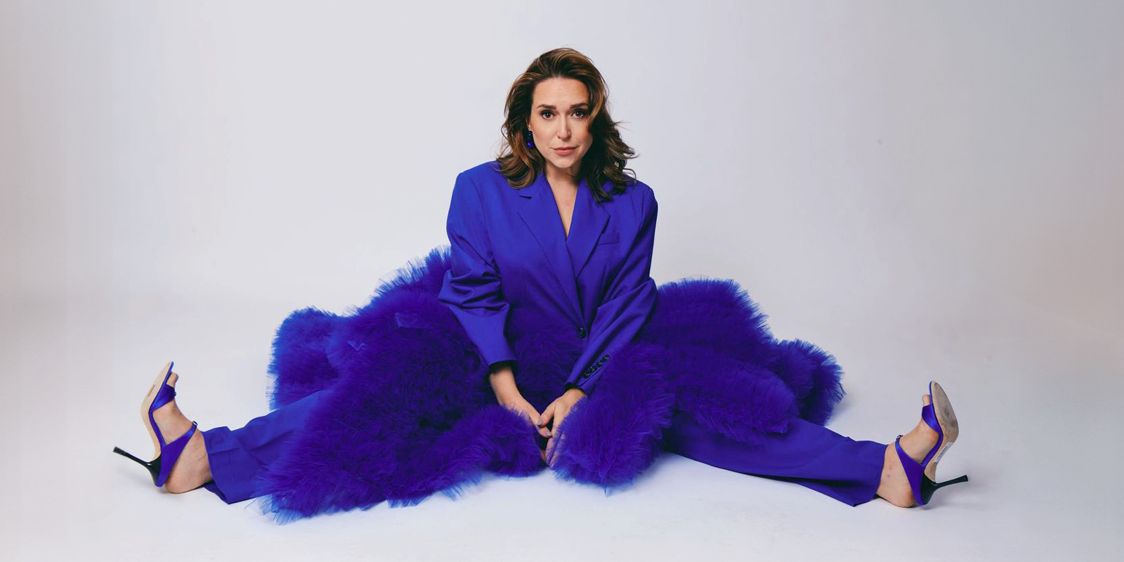 A woman in a periwinkle suit sits on a white floor wrapped in a purple boa.