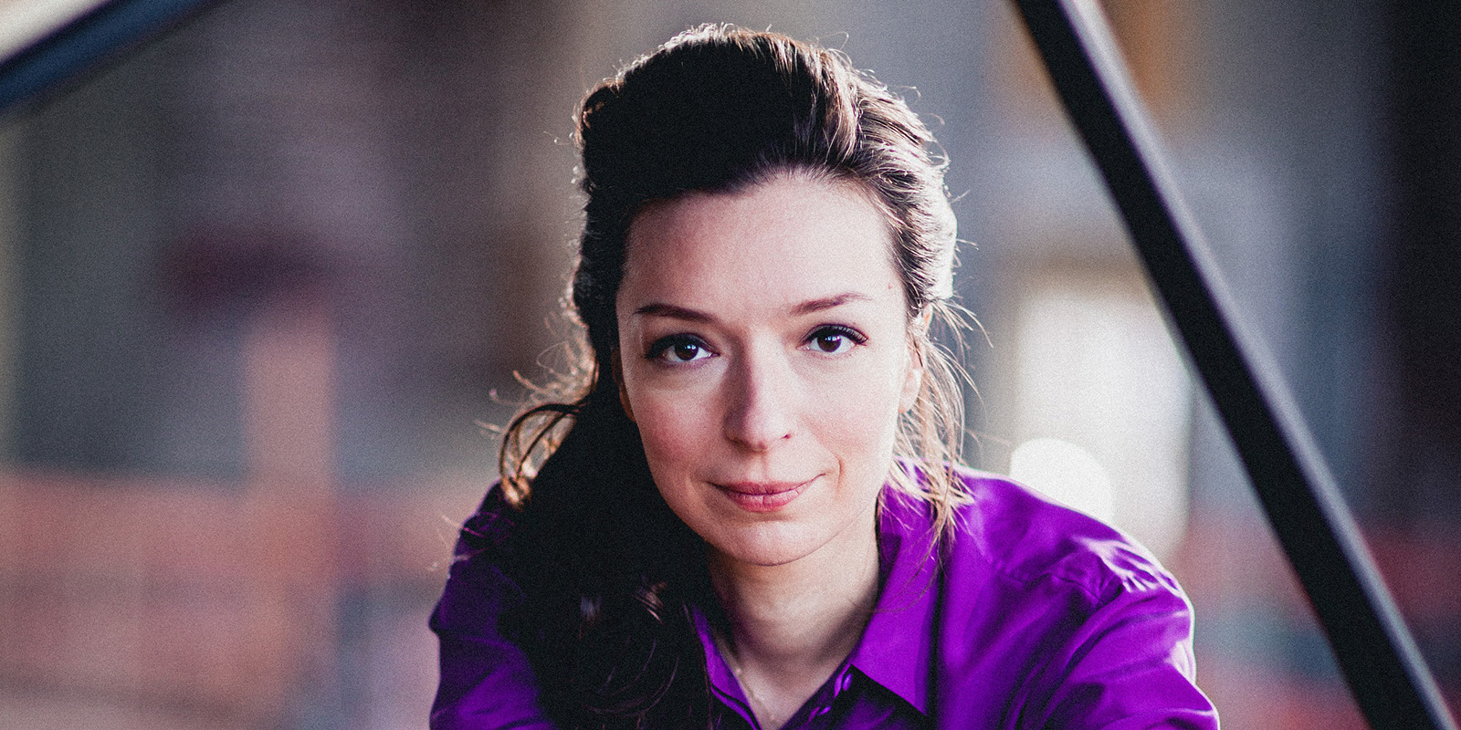 A woman with long brown hair wears a purple shirt and sits at a black piano.