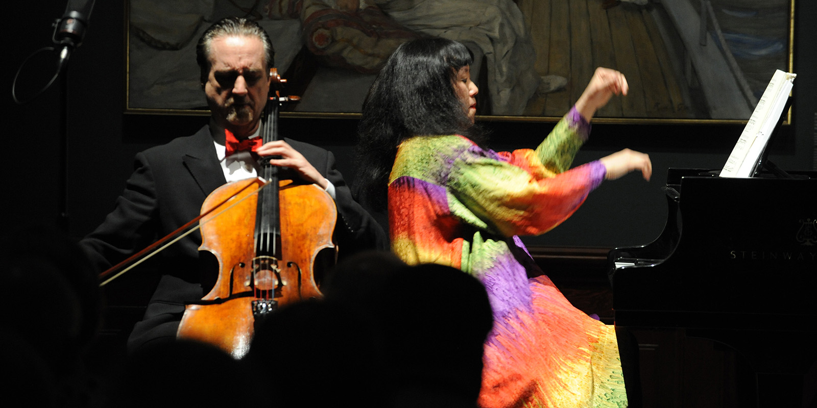 A man in a black tuxedo plays the cello and a woman in a multicolored dress plays the piano.