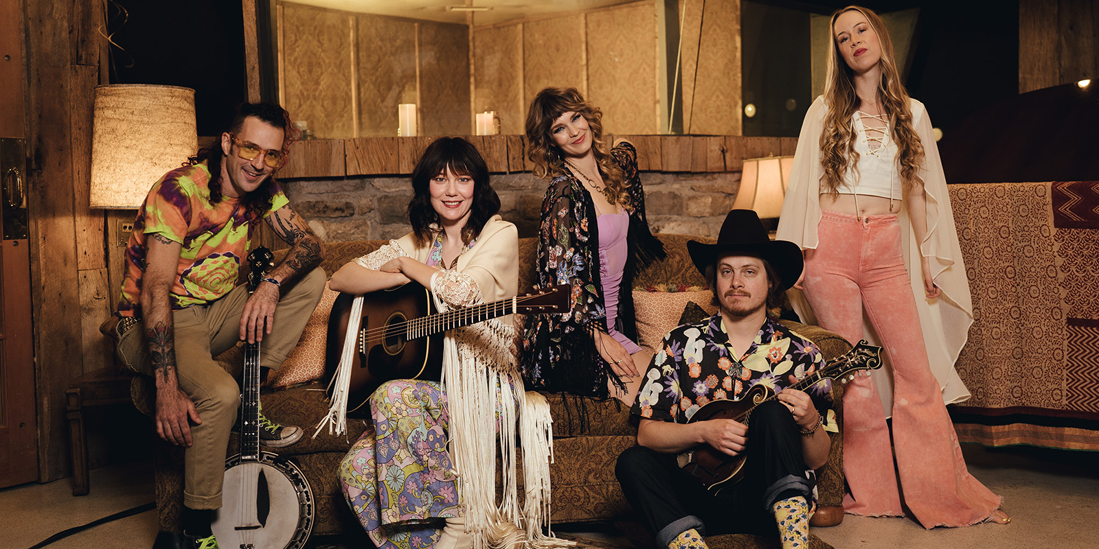 Five musicians sit and stand around a brown sofa in a wood paneled room.