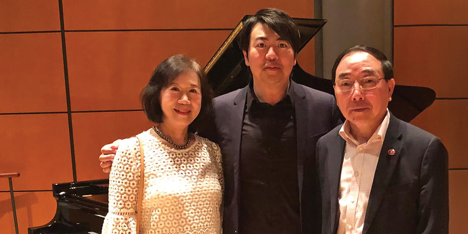 A woman and man stand on either side of the pianist Lang Lang in front of a piano.