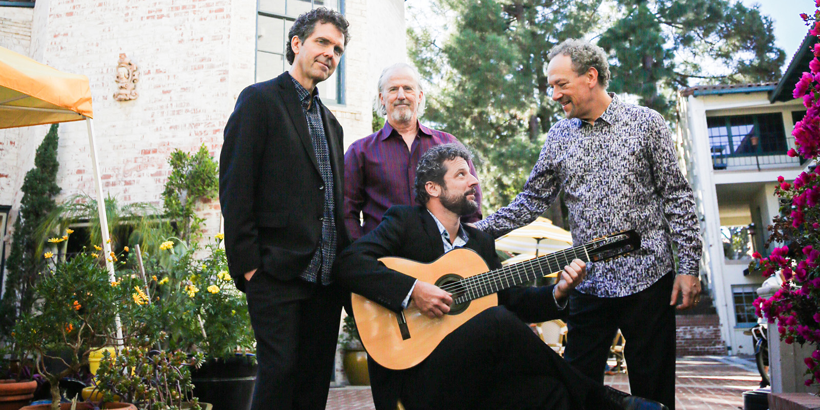 A man sits in a chair holding a guitar. Three other men stand around him.