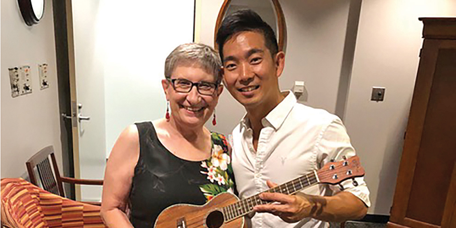 A woman in a black dress and a man in a white shirt stand close to each other and hold a ukulele.