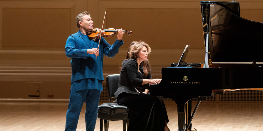 A man plays a violin next to a woman playing a piano on a stage.