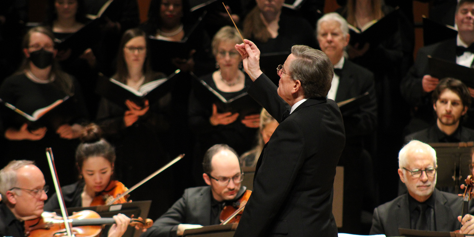 A man in a black suit conducts a choir and orchestra on stage.