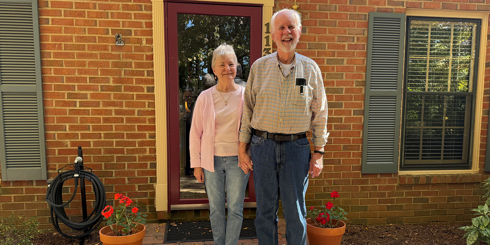 A woman and man stand close to each other in front of a red brick home.