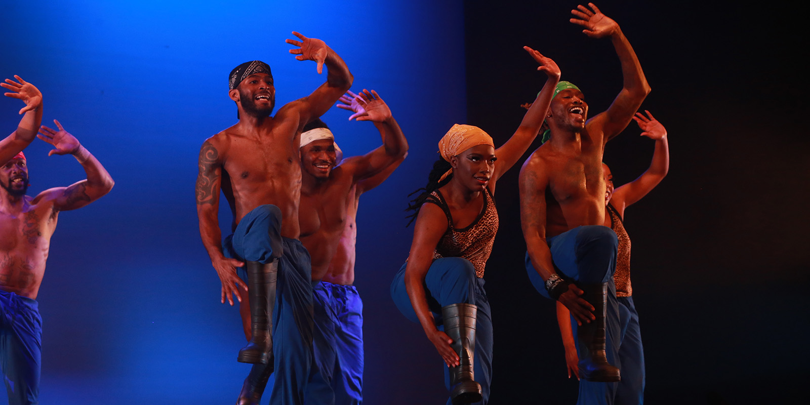 A group of dancers wear dark boots and vibrant blue pants on a stage in front of a blue backdrop.