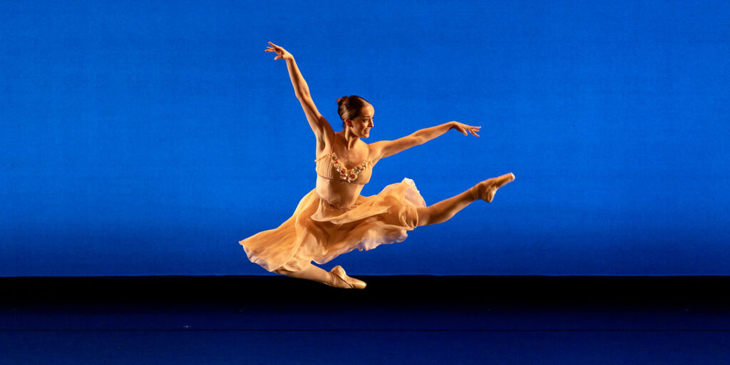 A woman in a peach colored dress leaps in the air on a stage in front of a blue backdrop.