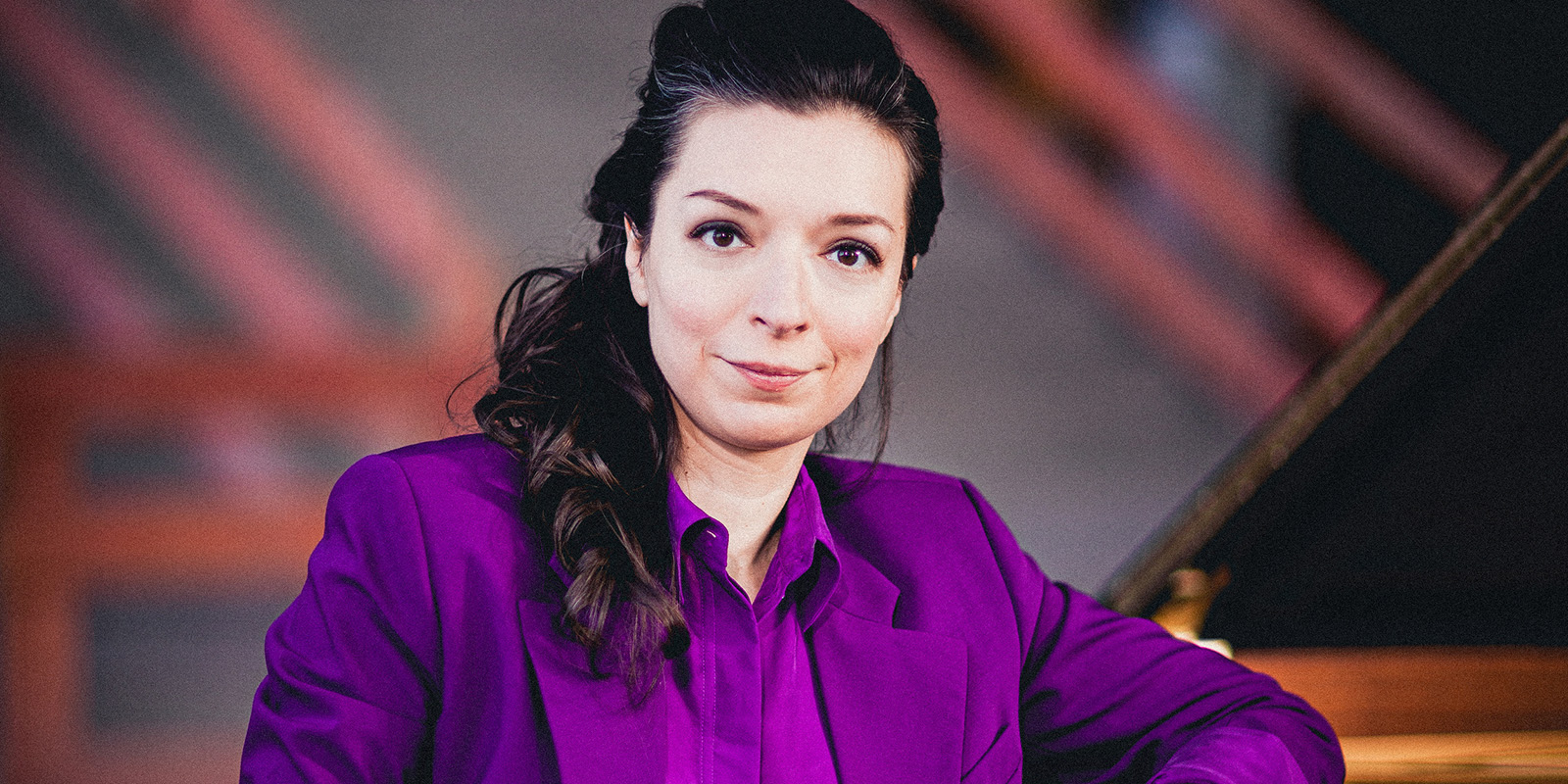 A woman with long, dark hair sits at a piano. She is wearing a purple shirt and jacket.