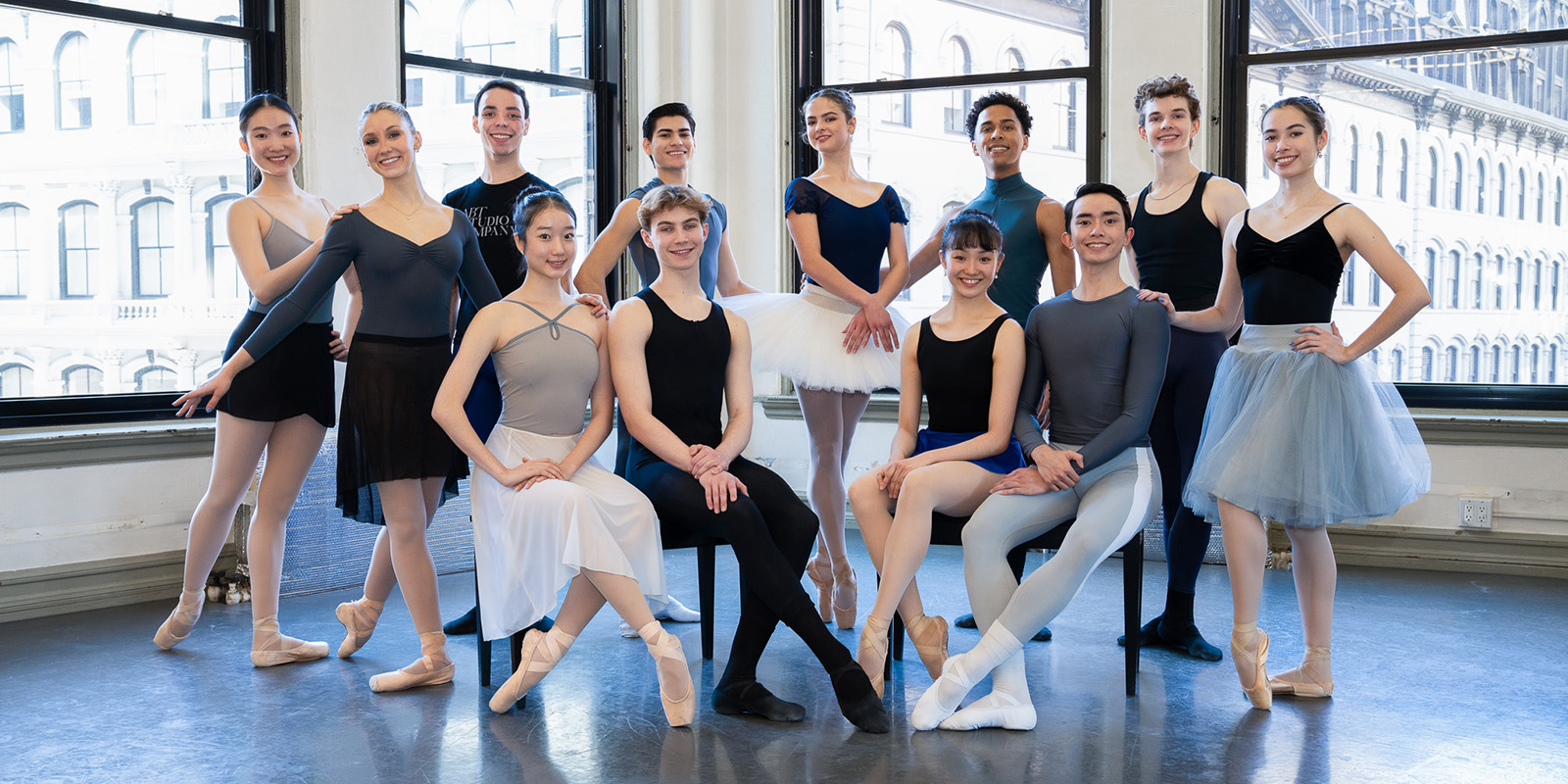 Young men and young women stand in a dance studio in front of large windows overlooking a city skyline.