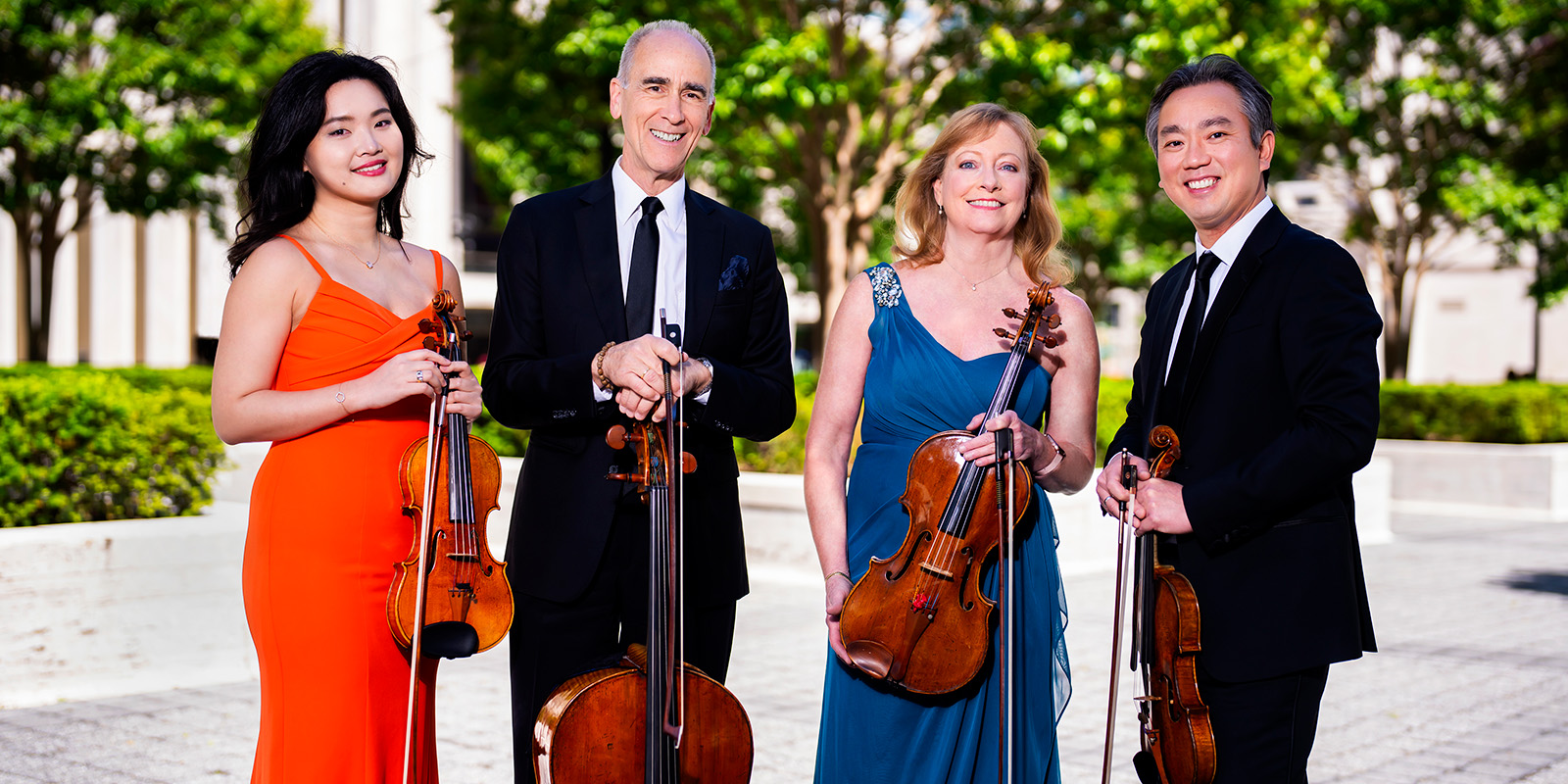 Two women in colorful dresses and two men in dark suits hold violins, a viola, and a cello in a bright, outdoor plaza.