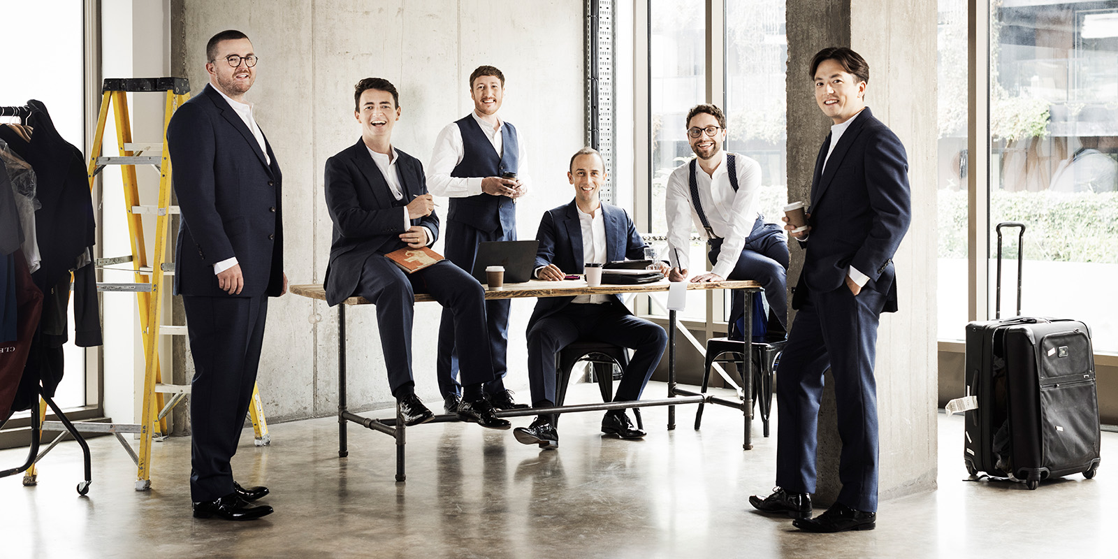 Six men in dark suits stand and sit in a studio with large windows.