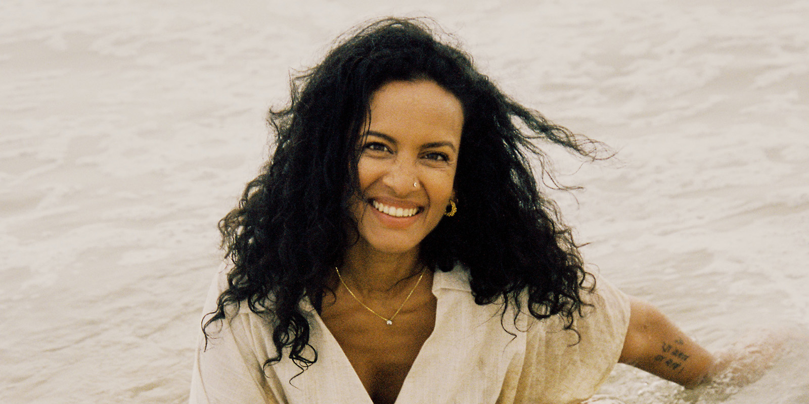 A woman in a white dress sits on the beach in front of the water.