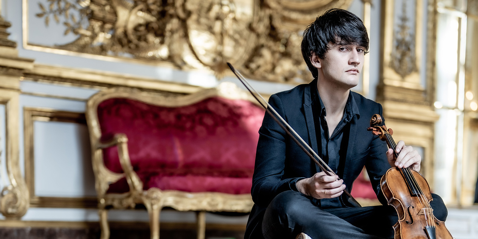 A man holding a violin sits on a floor in front of a red velvet sofa.