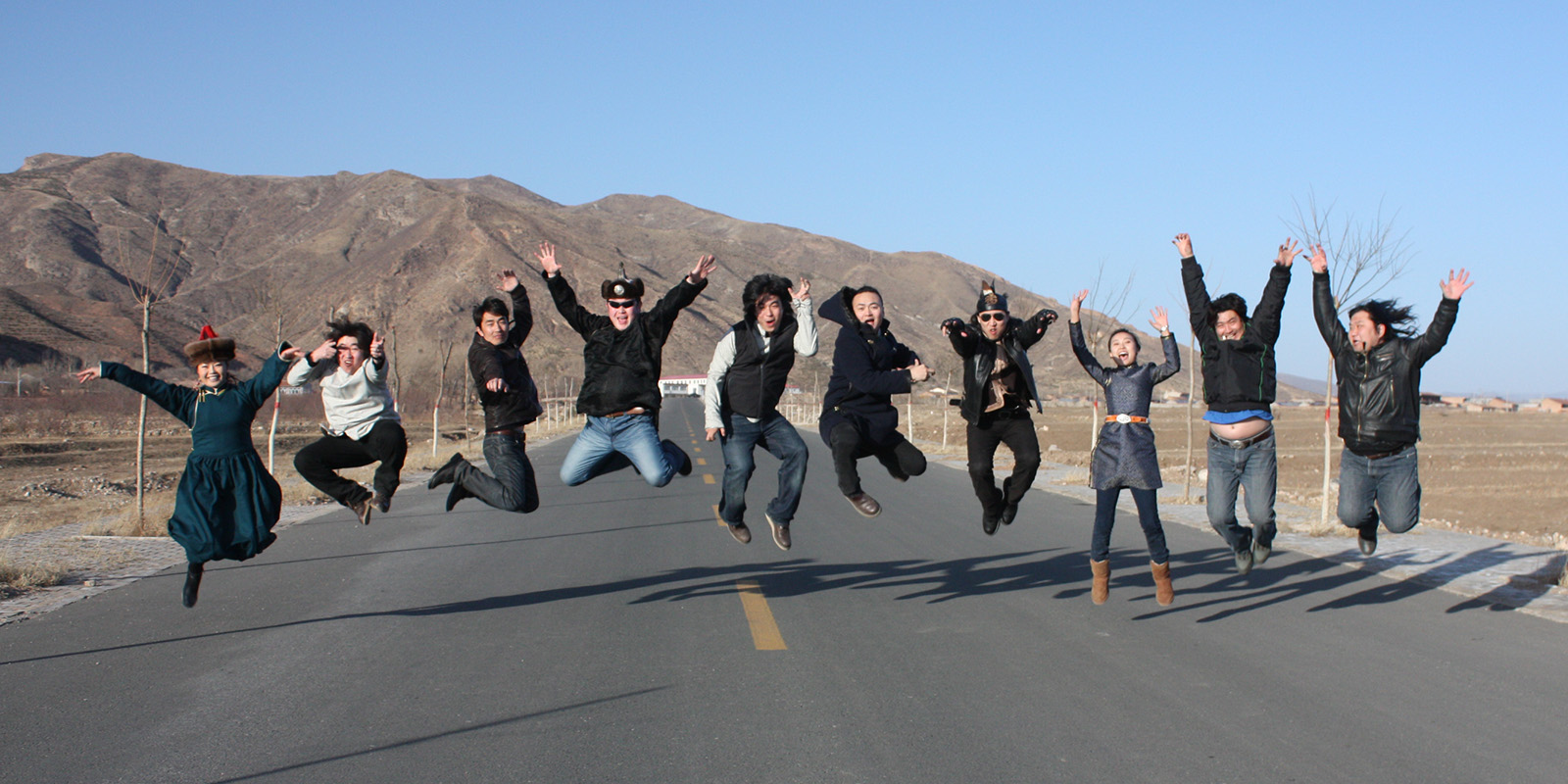 A group of men and women jump in the air above a road in a desert mountain area.