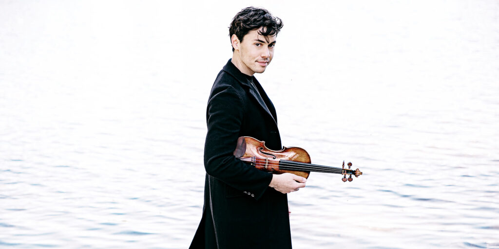 A man in a long, dark jacket stands in front of a body of water holding his violin.