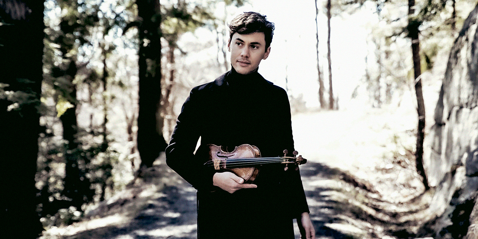 A young man in a dark coat stands on a forest path holding his violin.