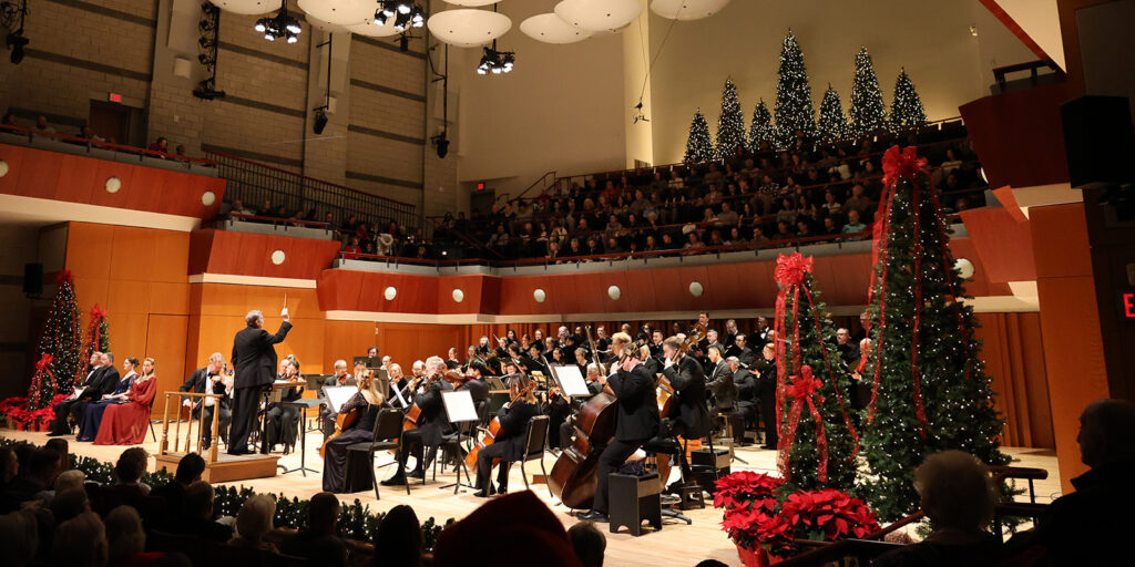 A conductor leads an orchestra in a festive concert hall decorated with Christmas trees, red ribbons, and poinsettias, while a full audience watches from tiered seating.