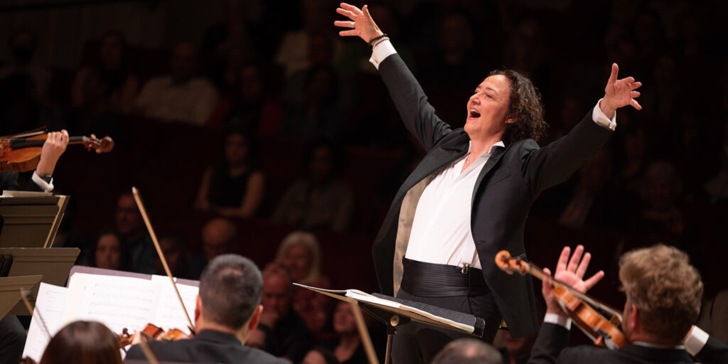 A conductor enthusiastically leads an orchestra, arms raised wide and expressive, while musicians play string instruments. The audience watches attentively in the background.