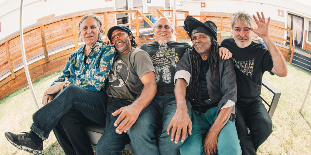 Five men sit closely together on an outdoor bench, smiling and laughing. Four have their arms around each other, and one man on the right waves at the camera. The setting is sunny, with wooden railings and grass in the background.