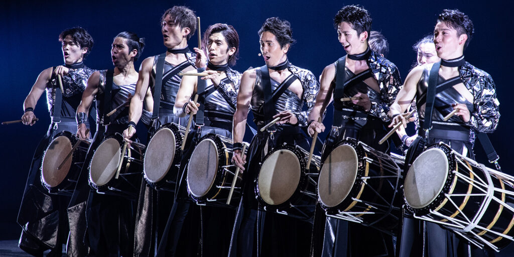 A group of eight performers in matching black and silver outfits play large traditional drums in unison on stage, under dramatic lighting.