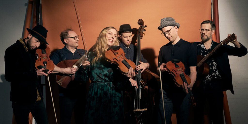 Six musicians stand together in front of an orange backdrop, each holding an instrument including violins, a double bass, and a guitar. A woman in a green dress smiles in the center while the others focus on their instruments.