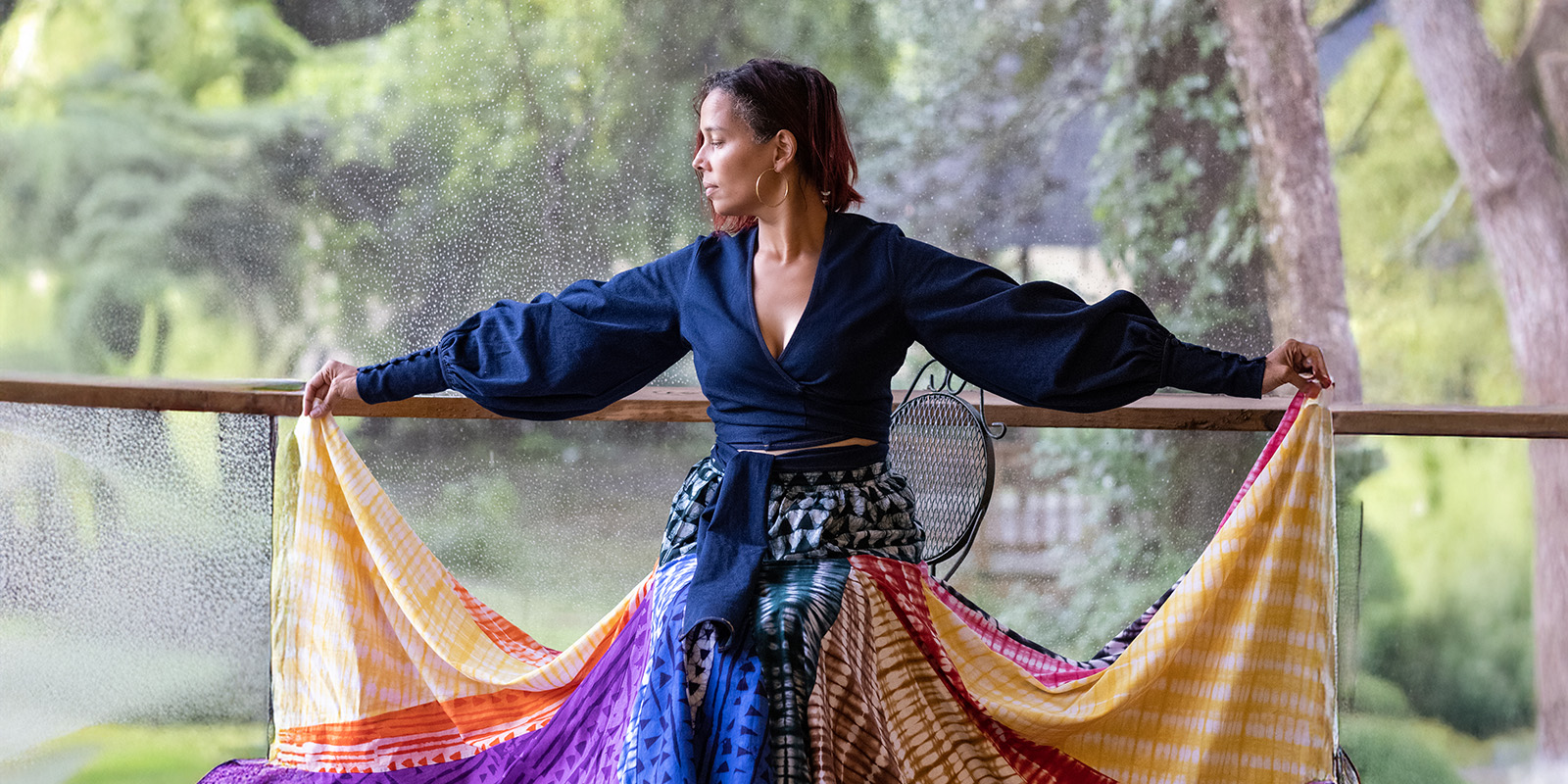 A woman stands on a porch holding out the colorful, flowing sides of her skirt. She wears a dark top with puffy sleeves and looks thoughtfully to the side. Trees and greenery are visible in the background.