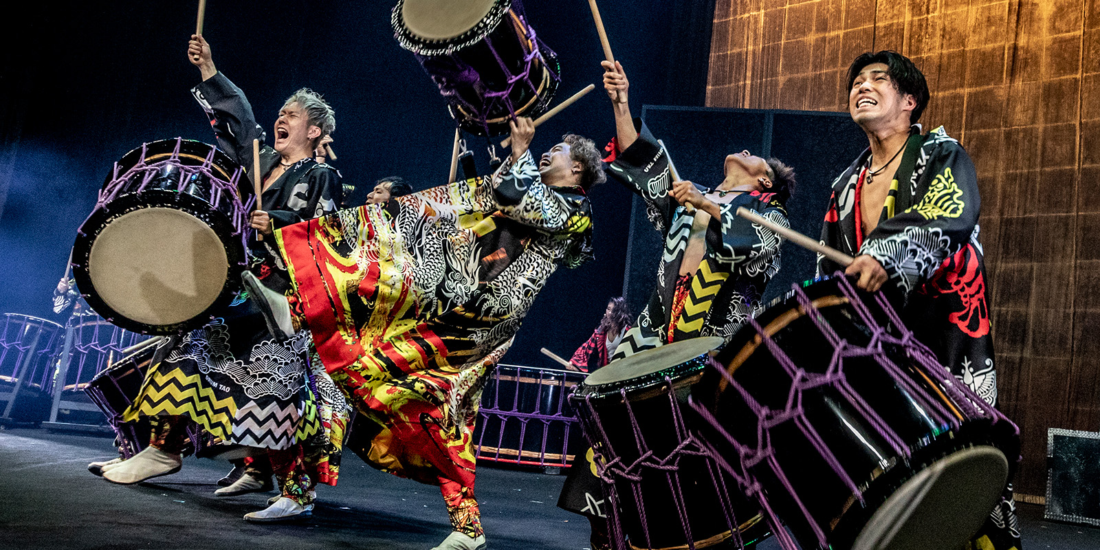 Four performers in vibrant traditional costumes energetically play large drums on stage, with expressive faces and dynamic poses, capturing the excitement and intensity of a live taiko drumming performance.