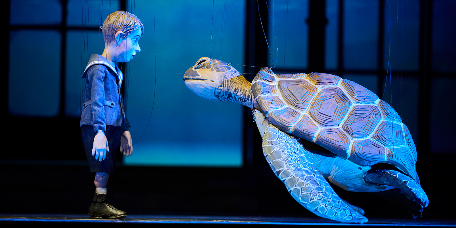 A boy puppet in a blue sailor suit stands facing a large puppet sea turtle, both suspended by strings on a dimly lit stage with a blue background.