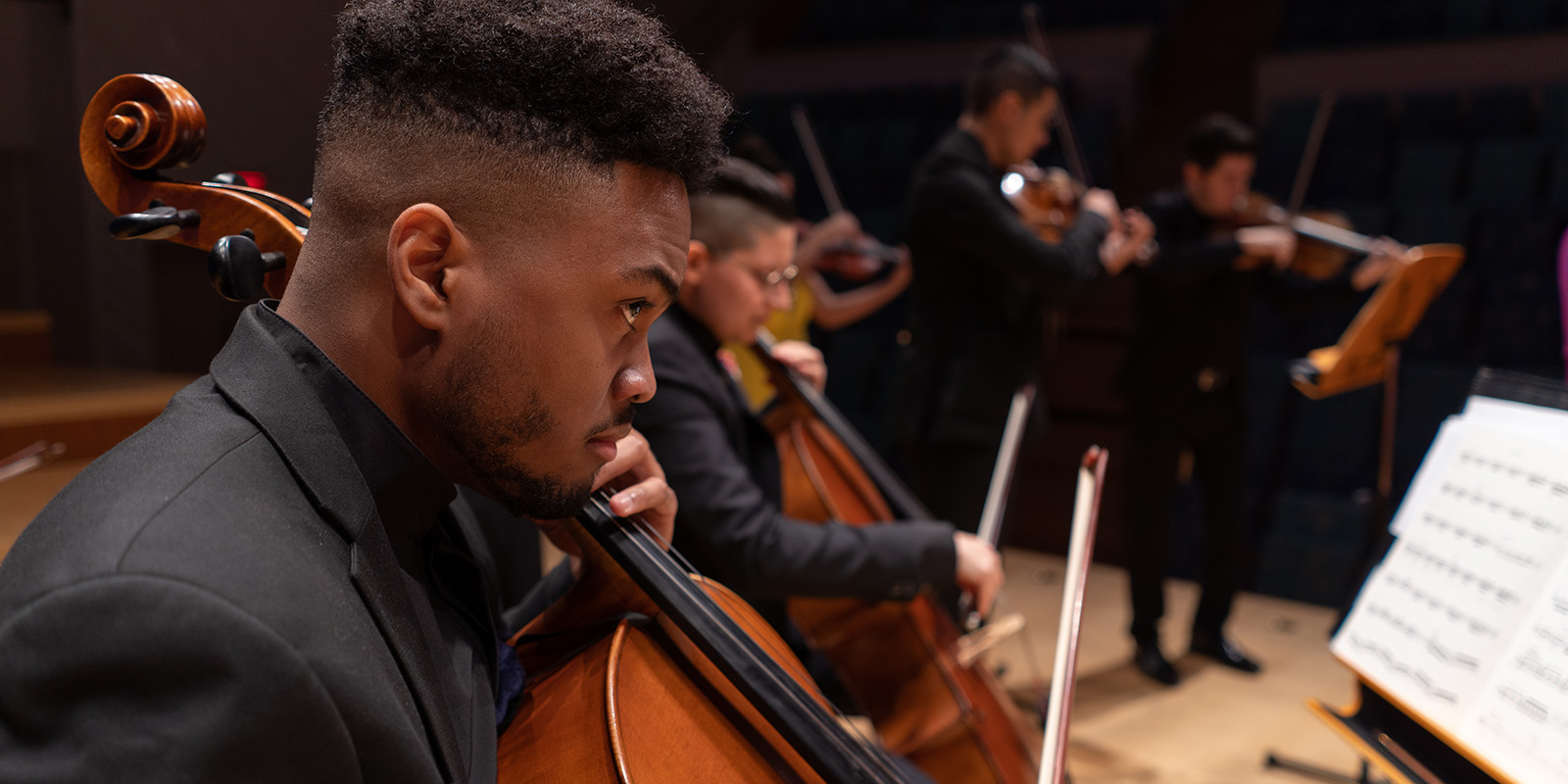 A group of musicians in formal black attire play string instruments, including cellos and violins, onstage in a concert hall, with sheet music visible in the foreground.