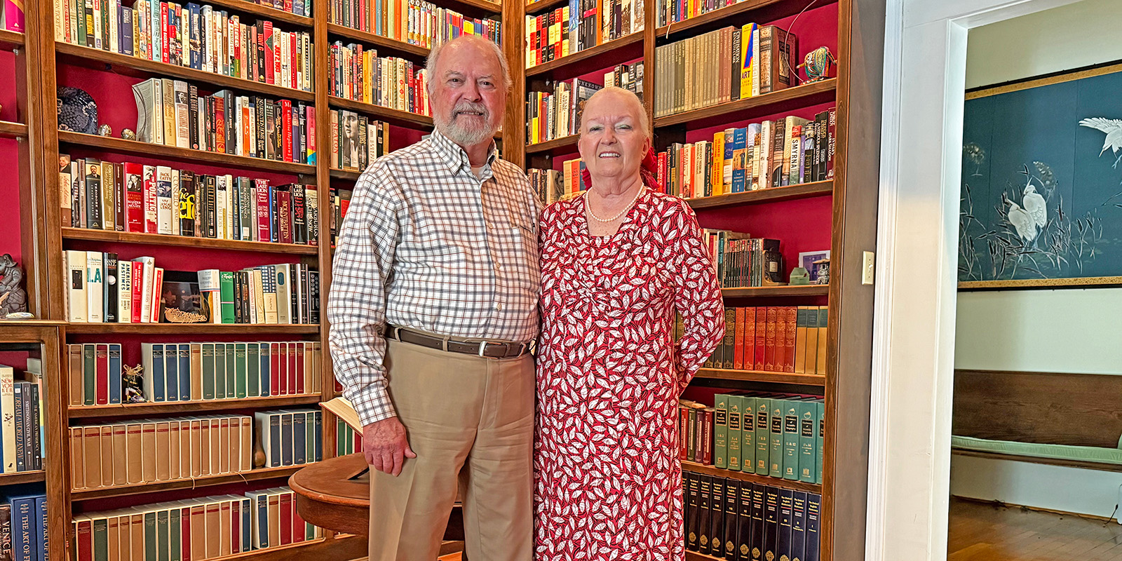 An older man and woman stand smiling together in a cozy library filled with bookshelves lined with colorful books. The woman wears a red and white patterned dress; the man wears a checked shirt and khaki pants.