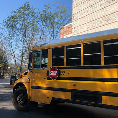 A yellow school bus with a visible stop sign and the number 22-2 is parked near a building, with trees and sunlight in the background.