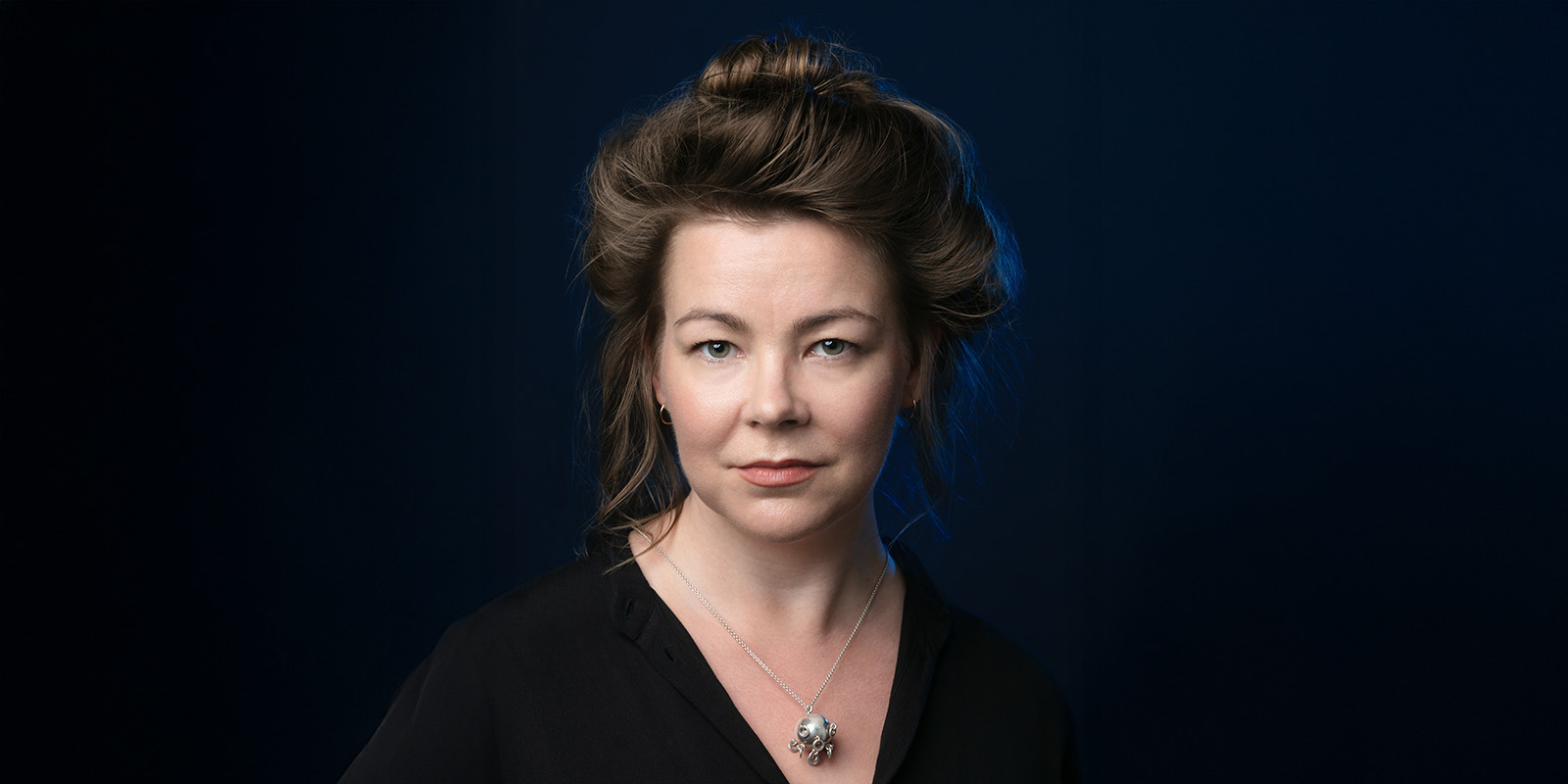 A woman with fair skin and brown hair styled in a loose updo, wearing a black top and a silver necklace, poses against a dark blue background, looking directly at the camera with a neutral expression.