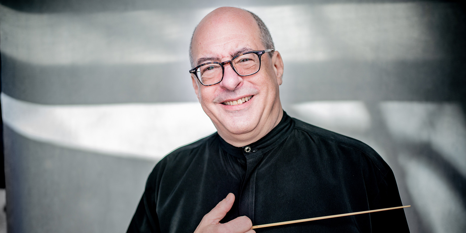 A smiling older man with glasses, wearing a black shirt, holds a conductors baton against his chest while standing in front of a softly lit, gray background.