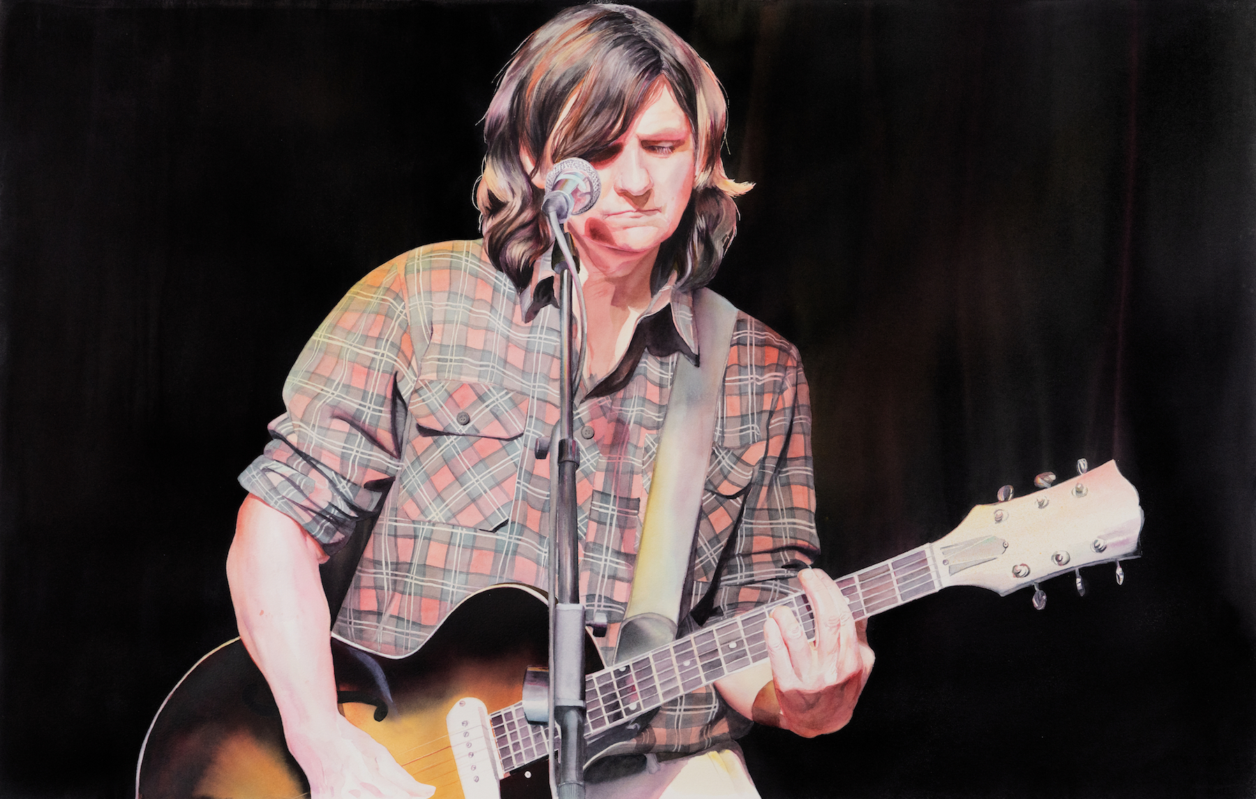 A person with long brown hair plays an electric guitar and sings into a microphone on stage, wearing a plaid shirt, with a dark background.