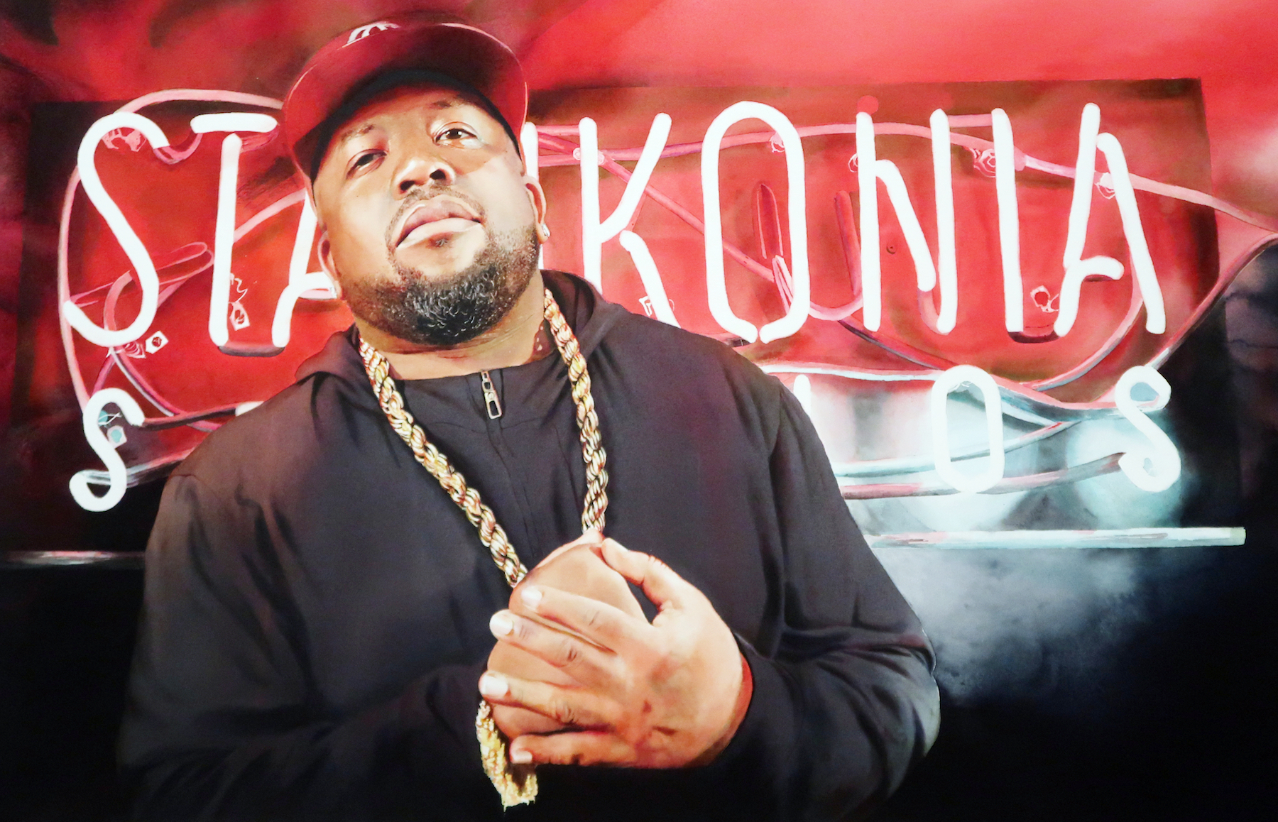 A man wearing a black hoodie, red cap, and gold chain stands in front of a neon sign that reads “Stankonia Studios” with red and black colors in the background.