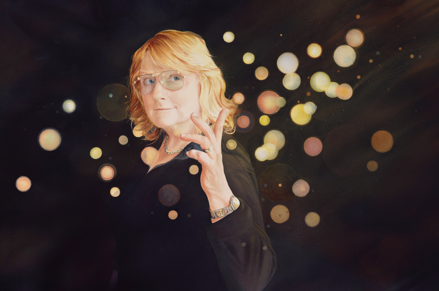 A person with shoulder-length blonde hair and glasses, dressed in black, stands against a dark background with glowing, out-of-focus light orbs, holding their hand up in a gesture.