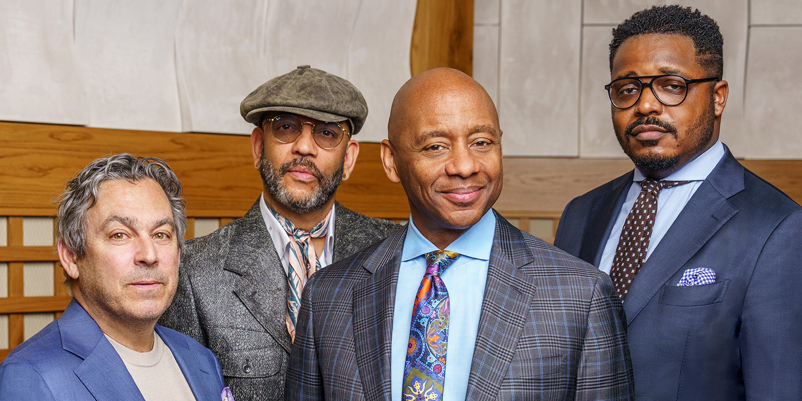 Four men in suits stand together indoors, posing for a group photo. The man in front center wears a plaid jacket and colorful tie; the others stand slightly behind him, all looking toward the camera.