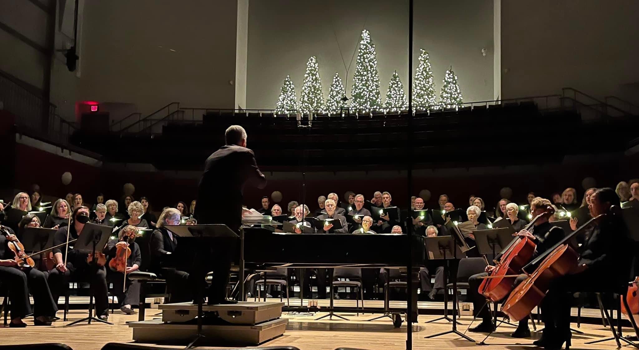 A conductor leads an orchestra and choir on stage, with string musicians seated in front and a choir behind. Lit, decorated trees are displayed at the back of the stage, suggesting a festive or holiday performance.