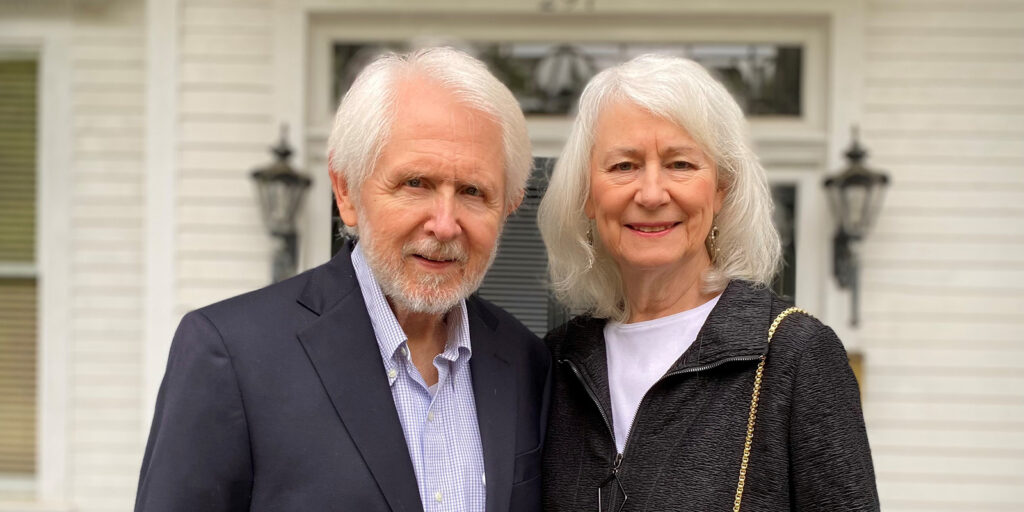 An older man in a suit jacket and an older woman in a black zip-up stand smiling together outside a white house with large windows and black lanterns on either side of the door.