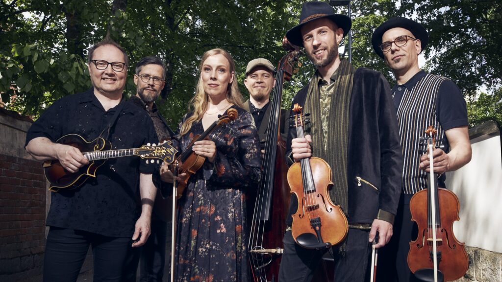 Six musicians stand outdoors, smiling and holding instruments including a mandolin, violin, upright bass, and guitar, with green trees and a brick wall in the background.