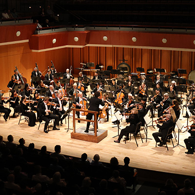 Atlanta Symphony Orchestra performs on stage in a concert hall, with musicians playing various instruments, a conductor leading from a podium, and an audience seated in front of the stage.