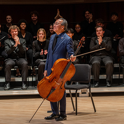 Yo-Yo Ma wears a blue suit and stands on stage holding his cello and bow, while an audience seated behind him claps and looks on appreciatively.