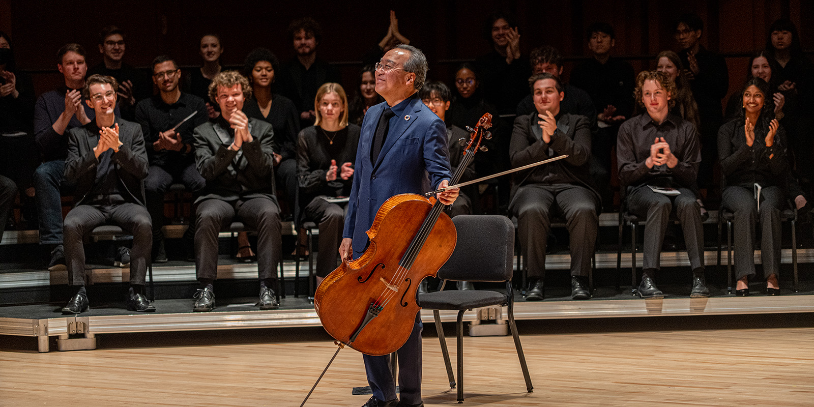 A cellist stands smiling and holding a cello and bow onstage, while an applauding audience and seated musicians wearing black attire are visible in the background.