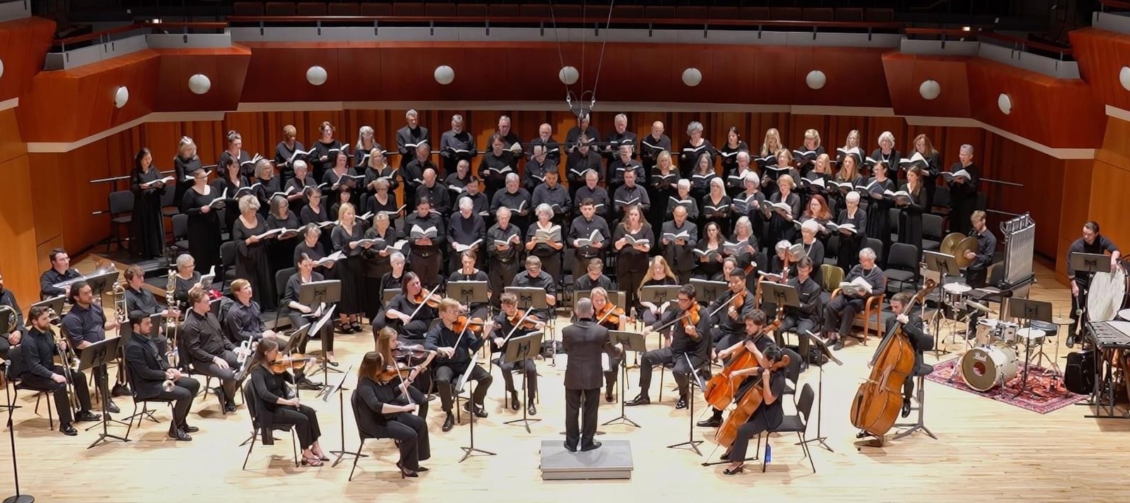 A large choir and orchestra perform on stage in a concert hall. The conductor stands in front, leading musicians playing string, brass, and percussion instruments, with choristers seated and singing behind them.